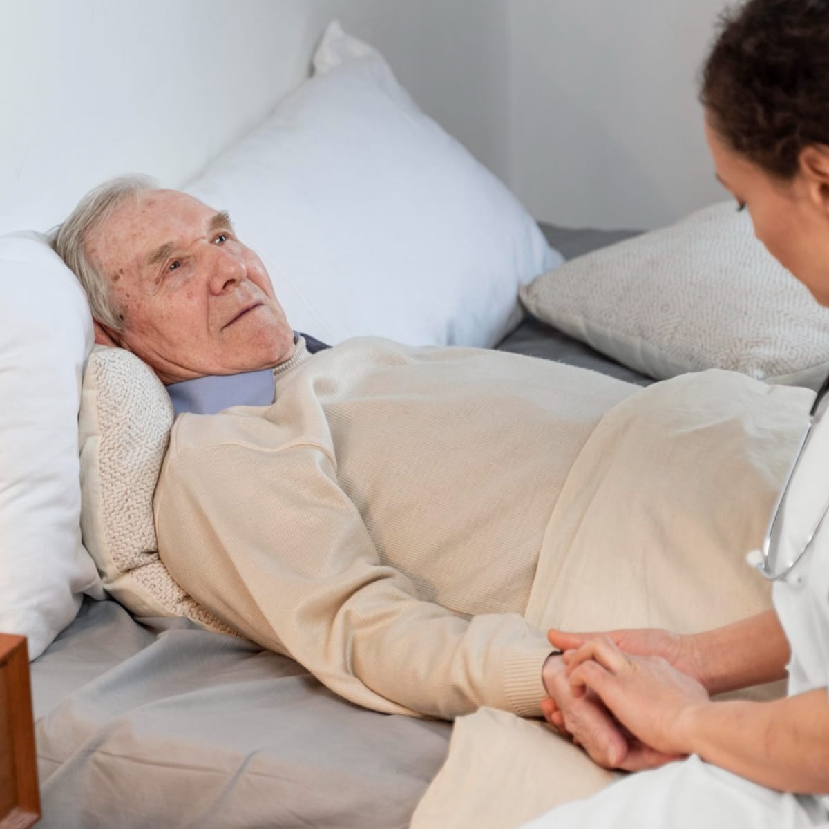 Elderly man laying in bed with a caregiver sitting at the edge of the bed